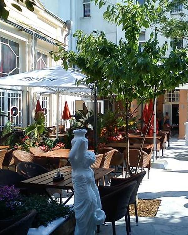 Gastgarten des Akropolis im begrünten Innenhof: Holztische unter Bäumen, rote Sonnenschirme und eine weiße Marmorstatue